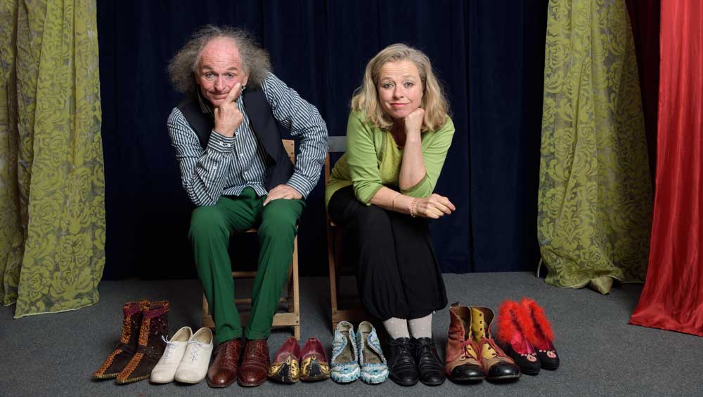 Mann und Frau sitzen auf Theaterbühne und haben acht Paar Schuhe vor sich.