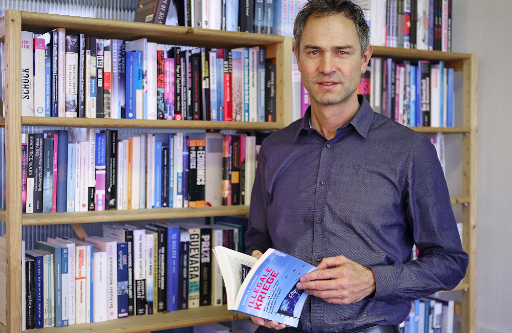 45-jähriger Mann mit grau melierten Haaren in graublauem Hemd steht vor einer Bücherwand und blickt in die Kamera