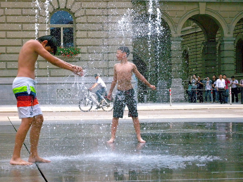 Zwei Jugendliche in Badeshorts auf dem Berner Bundeshausplatz, wo Wasserfontänen aus dem Boden schiessen