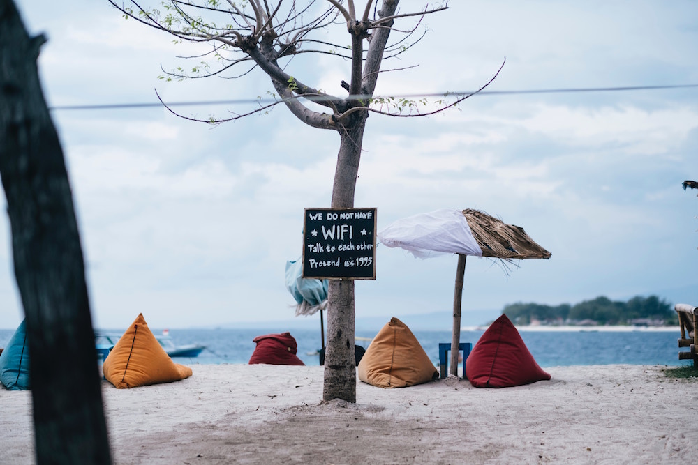 Bunte grosse Sitzkissen an einem Strand mit einem Schild am Baum, auf dem steht, dass es hier kein WLAN gibt und die Leute stattdessen miteinander reden sollen.