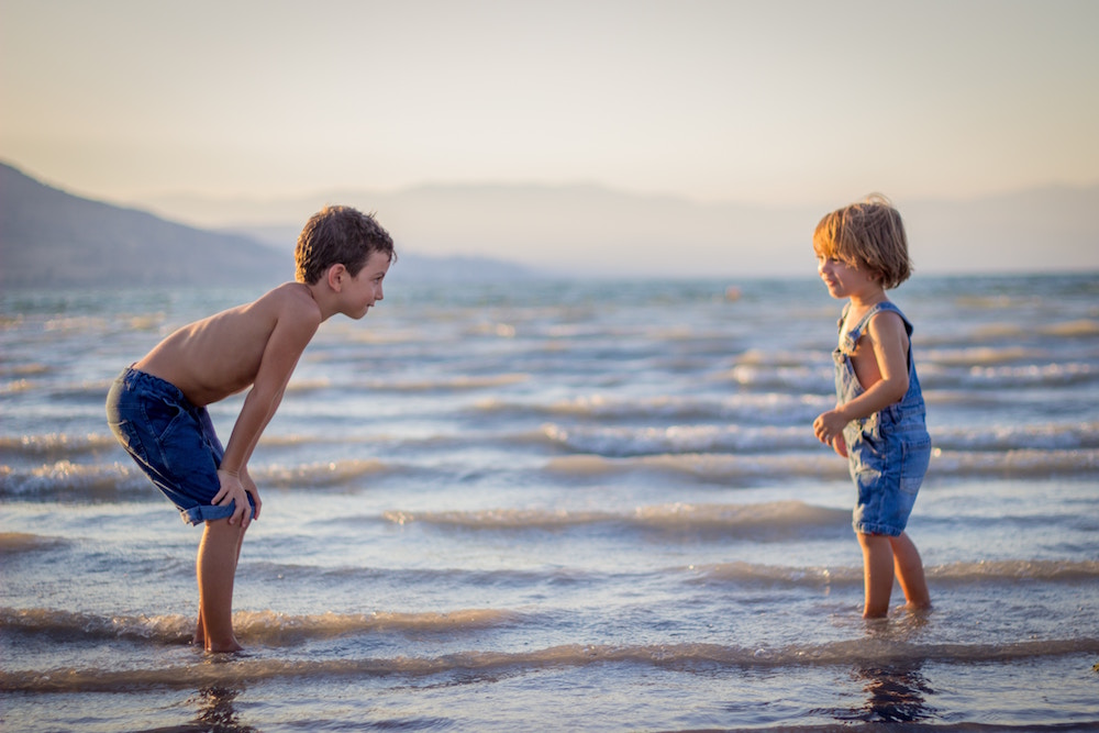 Zehnjähriger Junge und sechsjähriges Mädchen stehen mit den Füssen im Wasser am Strand und sehen sich freundlich an
