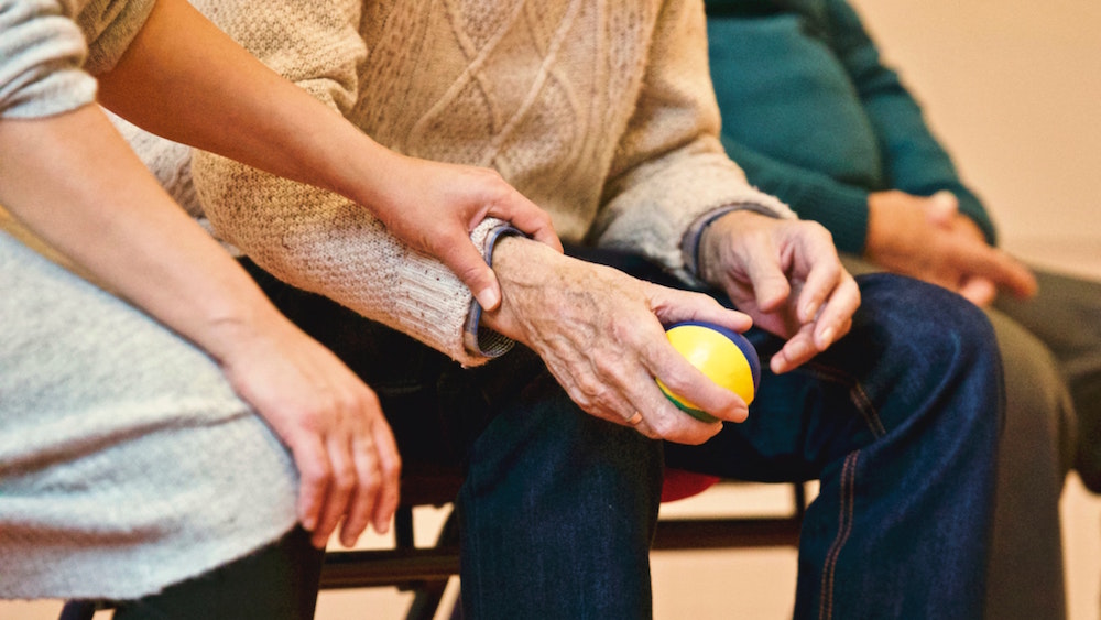 Alter Mann sitzt auf einem Stuhl, hält einen gelben Ball, eine Pflegeperson legt ihre Hand auf seinen Arm