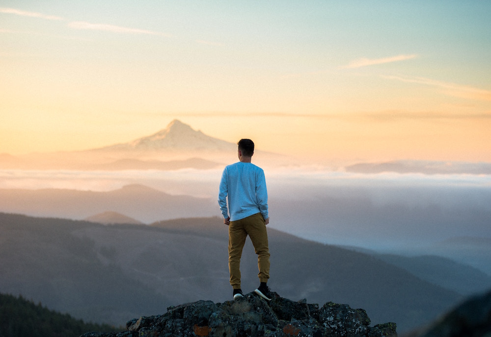 Junger Mann in hellblauem Pullover steht auf einem Felsen und blickt zu den Bergen in der Ferne