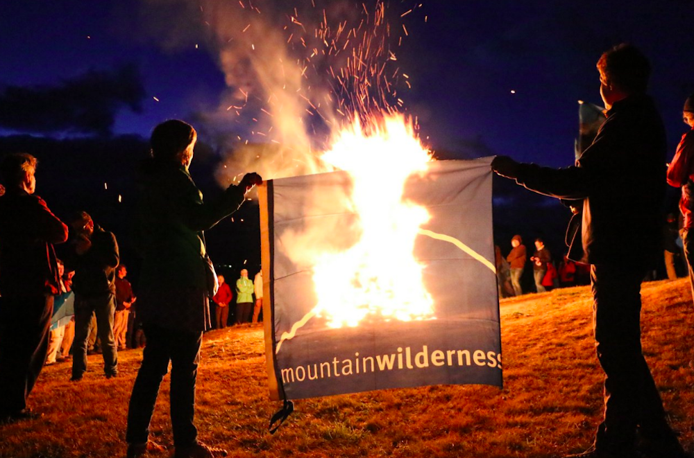 Mehrere Personen stehen in der Nacht um ein offenes Feuer auf einer Alpweide und protestieren gegen Heli-Skiing. Zwei halten eine Flagge mit der Aufschrift Mountain Wilderness