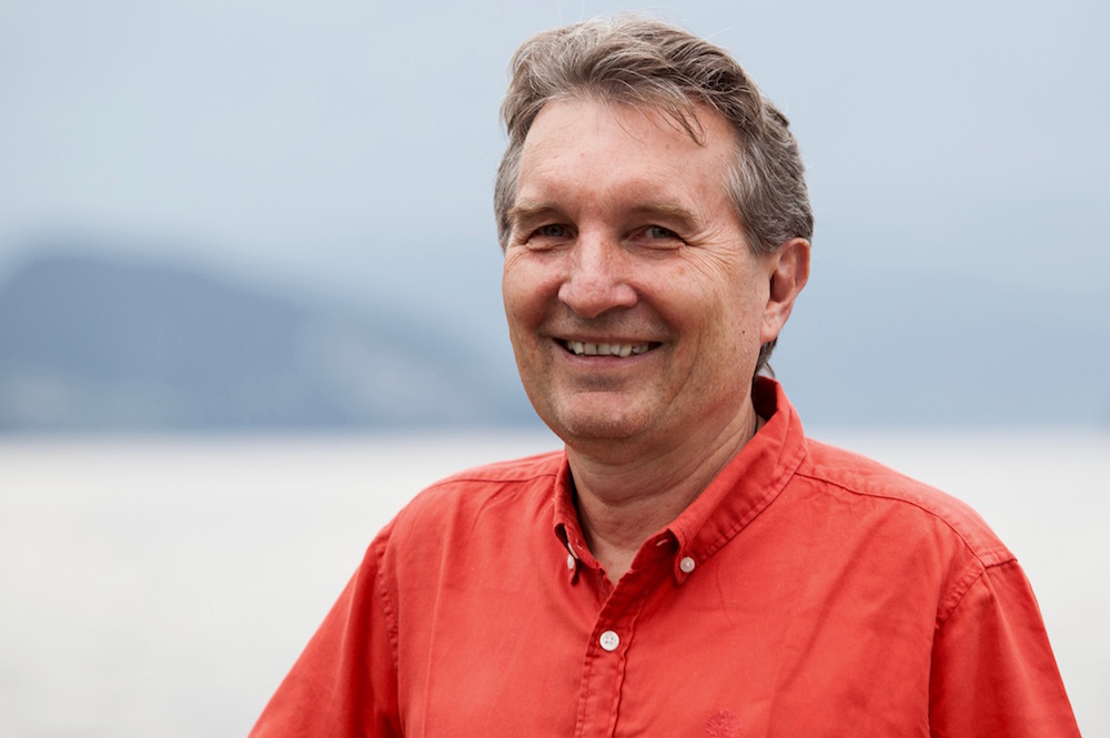 55-jähriger Mann mit rotem Hemd und kurzen Haaren blickt lächelnd in die Kamera, im Hintergrund See und Berge