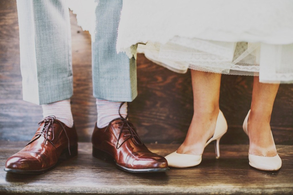 Ein Paar Männerbeine, Unterschenkel, braune Schuhe, weisse Socken, hellblaue Hose, und ein Paar Frauenbeine, weisse Schuhe mit Absatz, Stil 50er Jahre, nackte Füsse, Saum eines weissen Kleides, evtl. Hochzeitskleid
