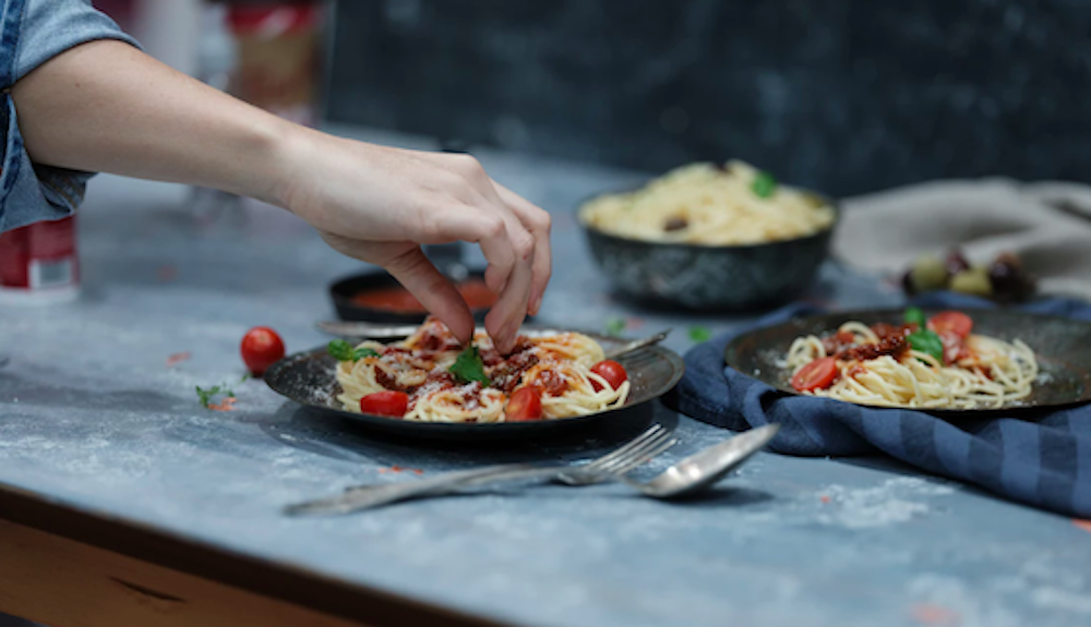 Eine Hand dekoriert einen Teller mit Spaghetti, Tomaten und Basilikum
