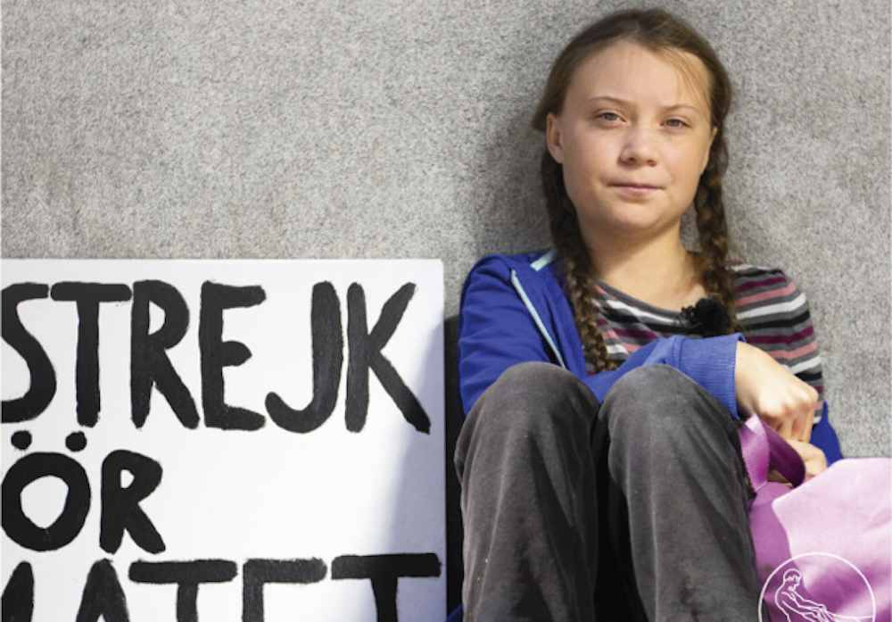 16-jähriges Mädchen mit braunen Zöpfen sitzt an eine Mauer gelehnt auf dem Boden, neben sich ein Schild, auf dem auf Schwedisch «Strejk» und weitere unvollständige Wörter stehen. Das Mädchen blickt in die Kamera