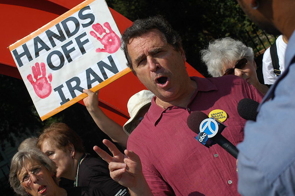 Blick in eine protestierende Menge, im Vordergrund ein Mann mit rotem Polo-Shirt, der etwas in ein Mikrofon spricht, und dahinter ein Schild mit der Aufschrift "Hands Off Iran".