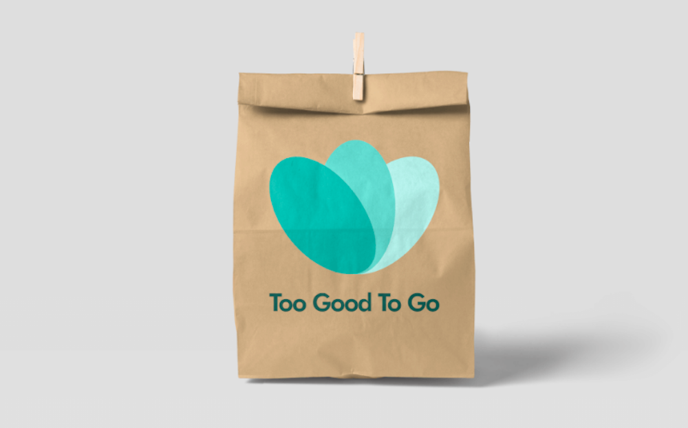 Braune Lebensmitteltüte mit dem Logo von Too Good To Go darauf, nämlich drei über einander liegenden ovalen Kreisen in drei Grünblau-Schattierungen