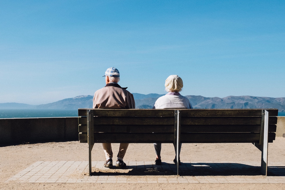 Älteres Paar sitzt mit dem Rücken zum Fotografen auf einer Bank auf einer Bergterrasse und betrachtet das Panorama